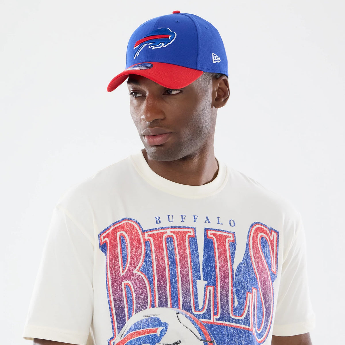 New Era Buffalo Bills NFL Sport Classic Cream T-Shirt, front view on a model wearing a blue and red cap, showcasing a large Bills graphic in blue and red letters on the chest with a distressed look and a detailed football image below.