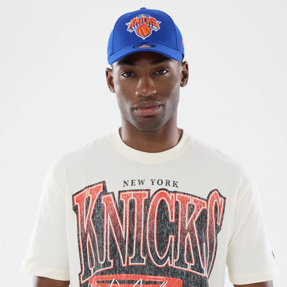 New Era New York Knicks NBA Sport Classic Cream T-Shirt, front view on a model styled with a blue cap, featuring large black and red Knicks lettering across the chest, New York text above, and a vintage effect graphic that adds to the retro style.