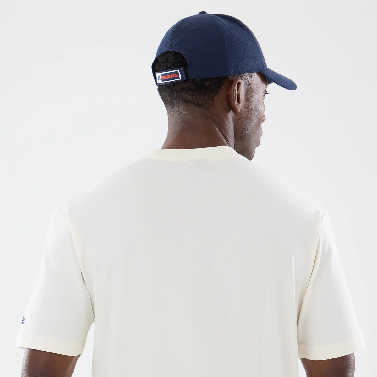 New Era Chicago Bears NFL Sport Classic Cream T-Shirt, back view on a model wearing a navy blue cap, showcasing the plain cream back of the shirt with stitching on the sleeves and a small embroidered Bears logo patch on the left sleeve.