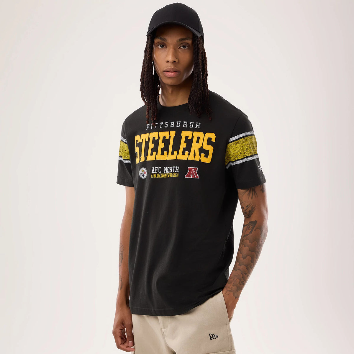 New Era Pittsburgh Steelers NFL Sideline 2026 Black T-Shirt, front view on a model wearing a black cap, features large yellow printed STEELERS lettering across the chest with team logos beneath and distinctive yellow and white stripes on the sleeves.