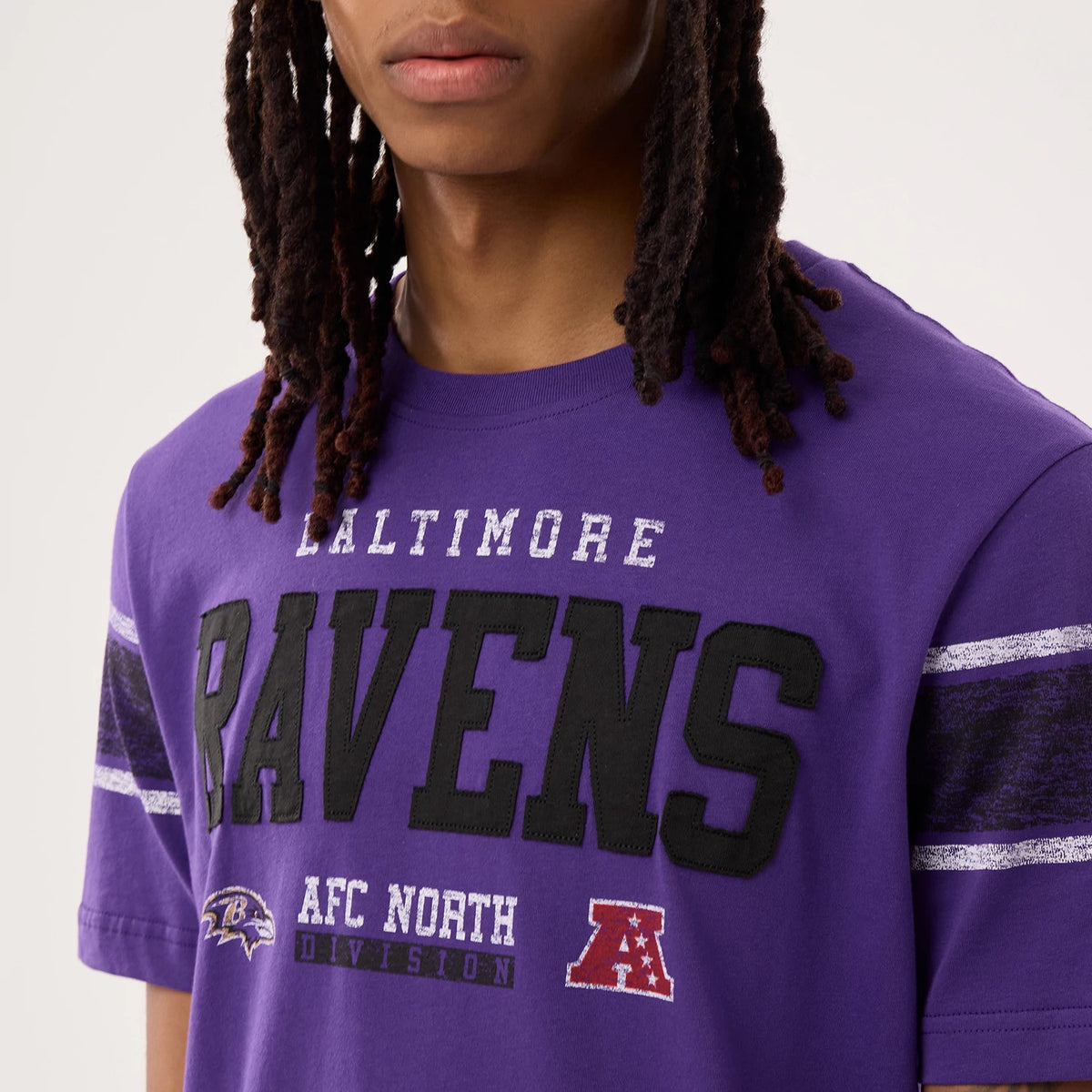New Era Baltimore Ravens NFL Sideline 2026 Purple T-Shirt, front view on a model showing black embroidered Ravens lettering across the chest, Baltimore text above, AFC North Division text below, and team logos printed near the bottom of the shirt.