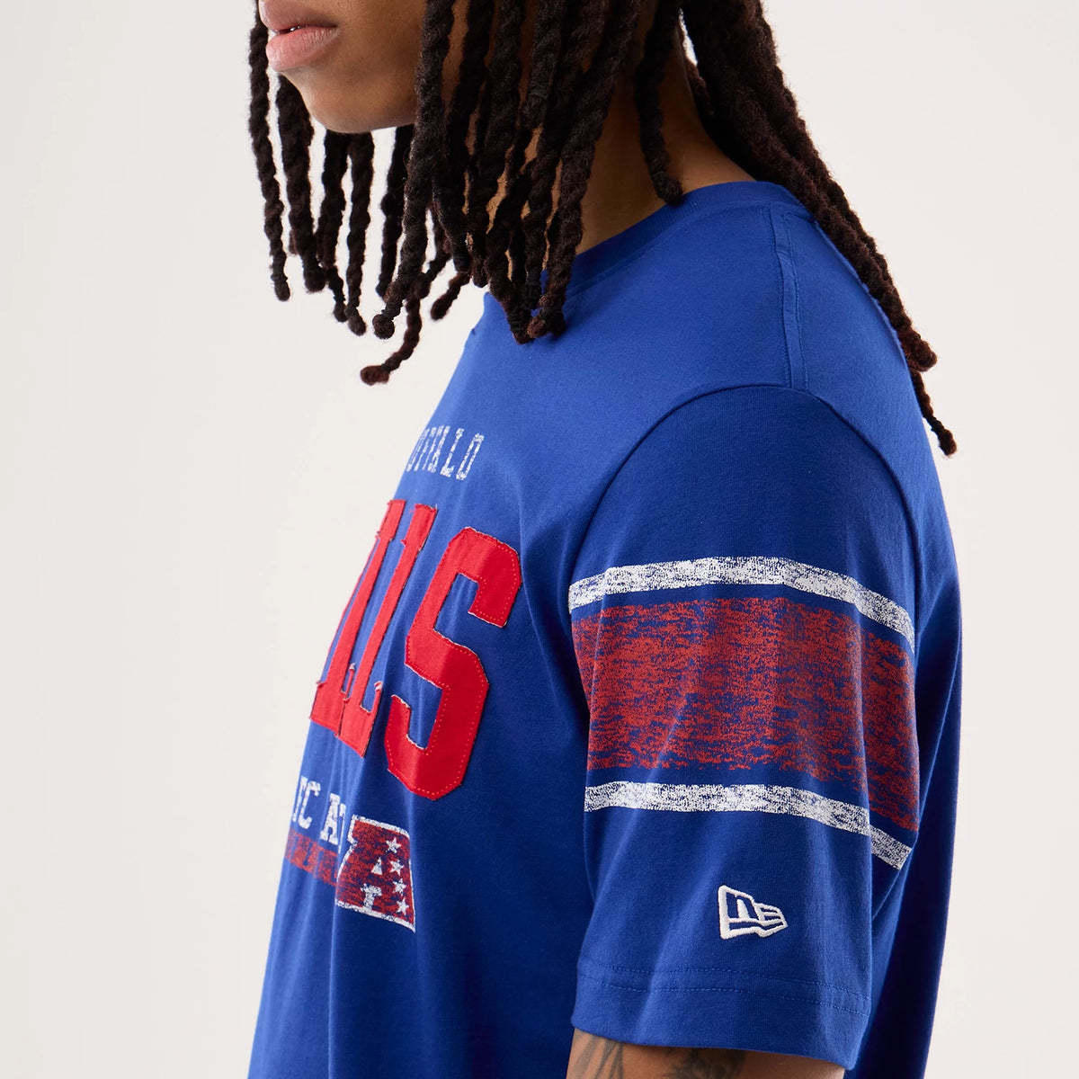 New Era Buffalo Bills NFL Sideline 2026 Blue T-Shirt, side view on-model showing a red and white graphic spell-out across the front, distressed red and white stripes on the sleeve, and a white embroidered New Era logo near the hem.