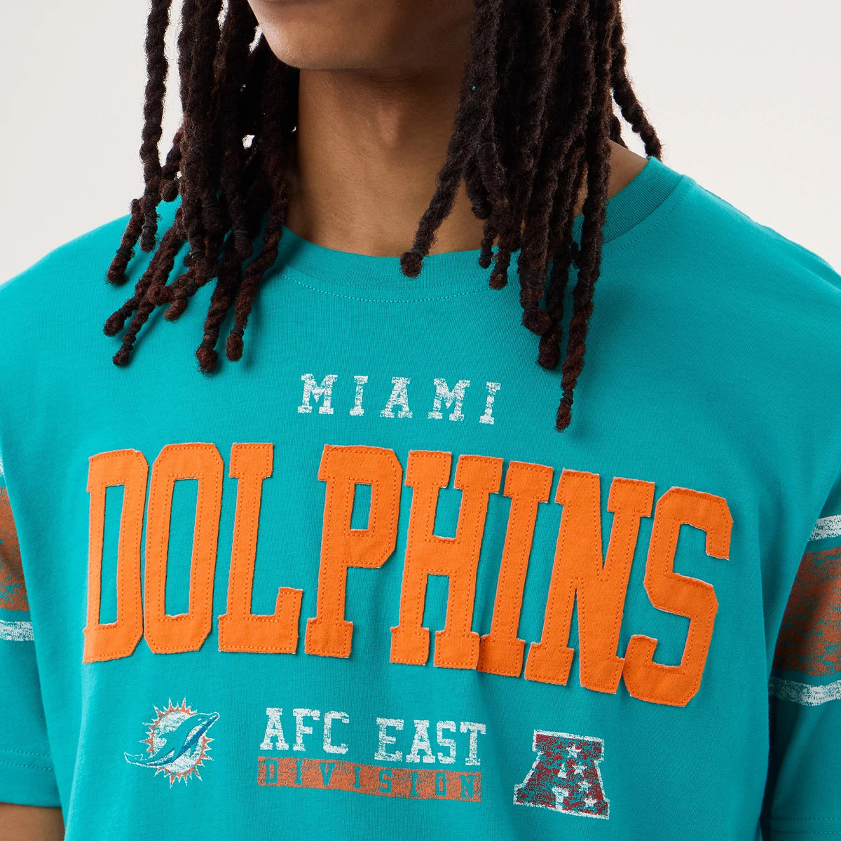 New Era Miami Dolphins NFL Sideline 2026 Turquoise T-Shirt, front view on-model showing large orange Dolphins lettering across the chest, a white embroidered Miami above, and teal and orange AFC East Division logos below with striped sleeve details.