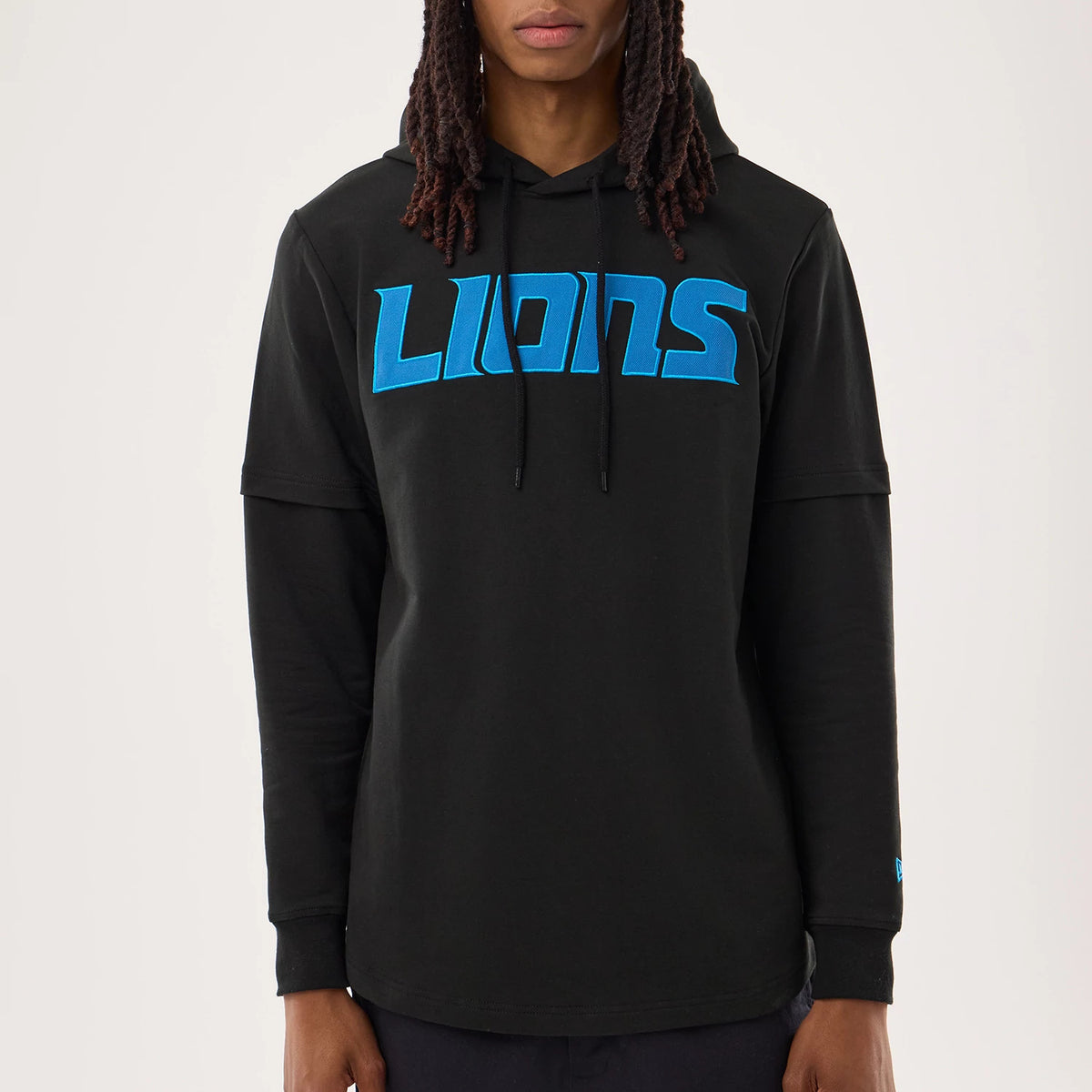 New Era Detroit Lions NFL Sideline 2026 Black Double Sleeve Hoodie, front view on a model showing the blue embroidered LIONS lettering across the chest, a black drawstring hood, and a small embroidered New Era logo near the left wrist.