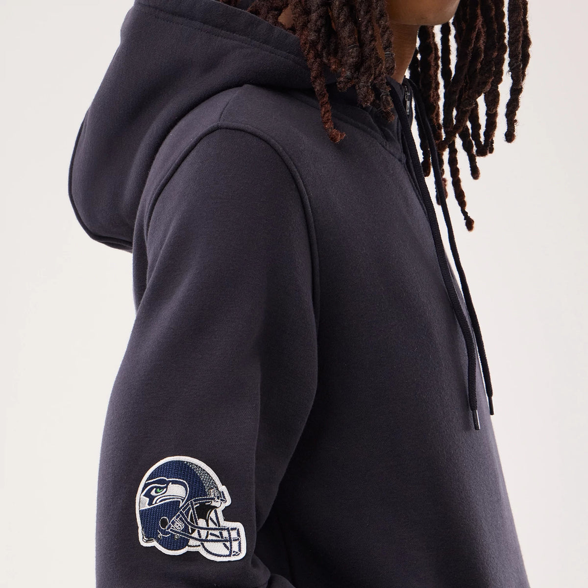 New Era Seattle Seahawks NFL Sideline 2026 Navy Pullover Hoodie, side view with the embroidered Seahawks helmet logo on the left sleeve, the navy hood resting on the shoulders, and a glimpse of drawstring cord hanging in front.