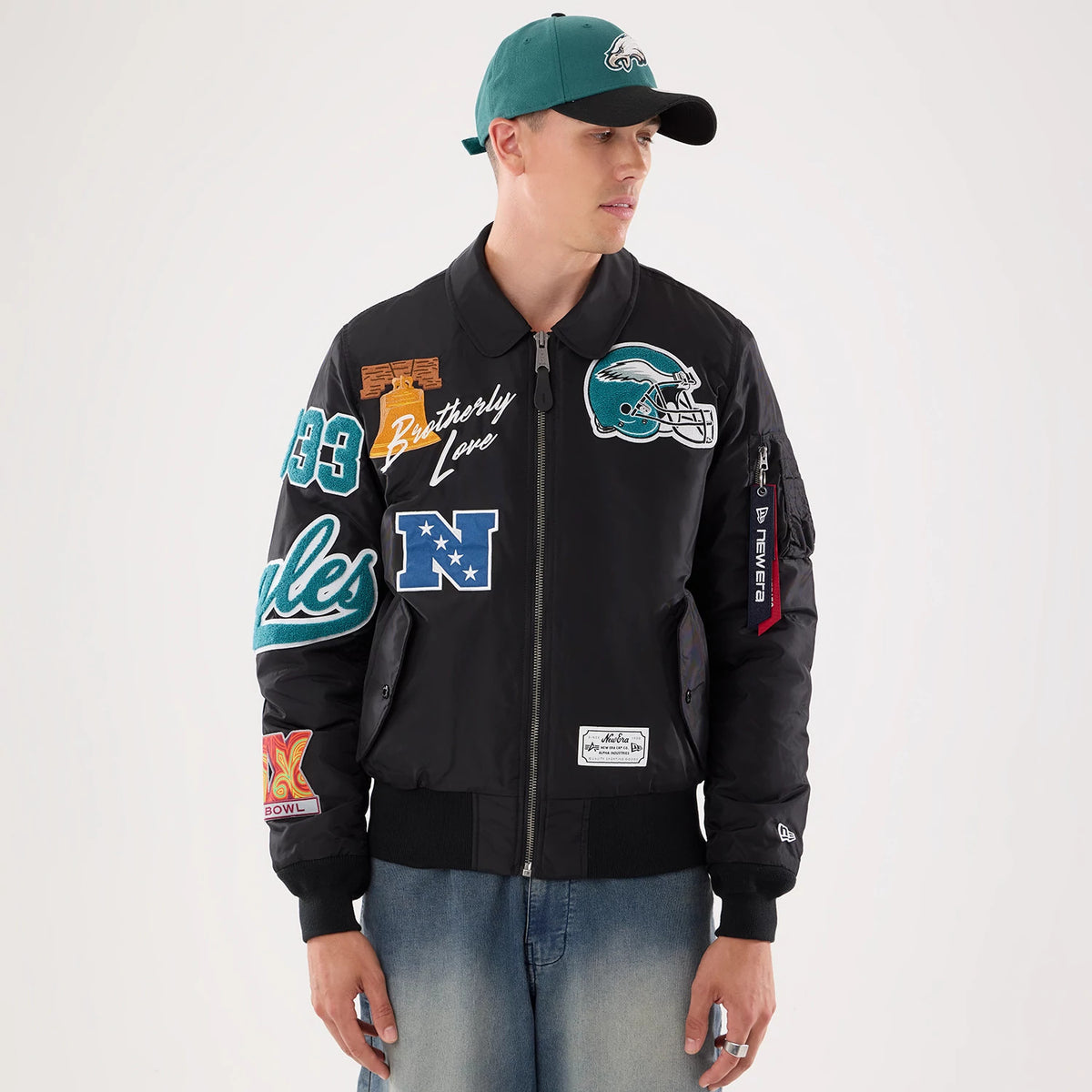 New Era Philadelphia Eagles NFL X Alpha Black Jacket, front view on-model showing multiple embroidered patches including a large Eagles logo on the chest, Brotherly Love script, and NFC emblem, with zipped arm pocket and New Era logo on left wrist.