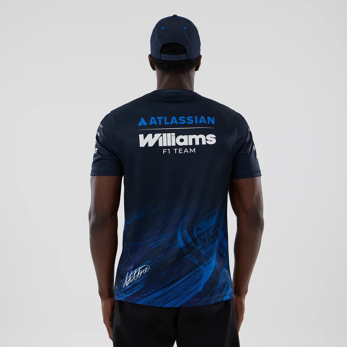New Era Williams Alex Albon Navy Tech T-shirt, back view on a model showing ATLASSIAN and Williams F1 Team lettering in white on the upper back, with a dynamic blue graphic pattern and an autograph detail near the base.