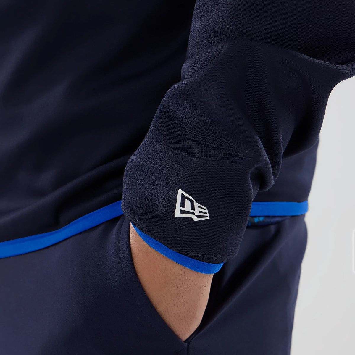 New Era Williams Navy Mid Layer 1/4 Zip Sweater, on-model close-up showing the New Era flag logo embroidered on the left sleeve near the cuff, with contrasting blue trim at the hem and sleeve, and a relaxed fit around the hand in the pocket.