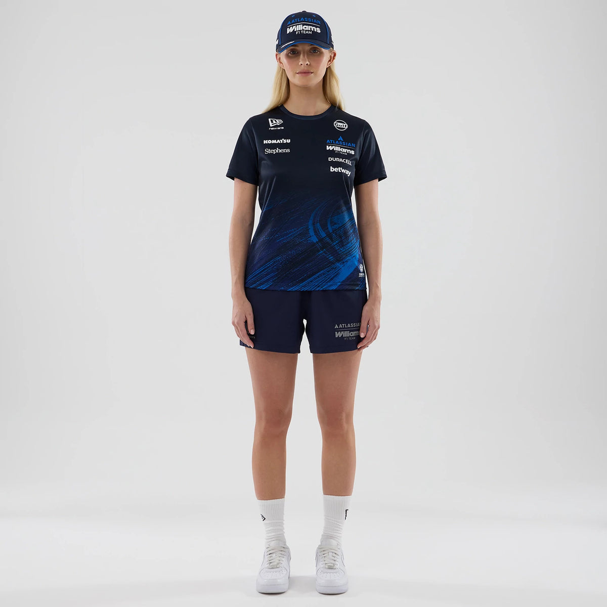 New Era Williams Womens Navy Tech T-shirt, front view on-model featuring a navy design with dynamic blue graphics on the lower half, various white sponsor logos across the chest, and a New Era logo on the sleeve; styled with matching shorts and cap.