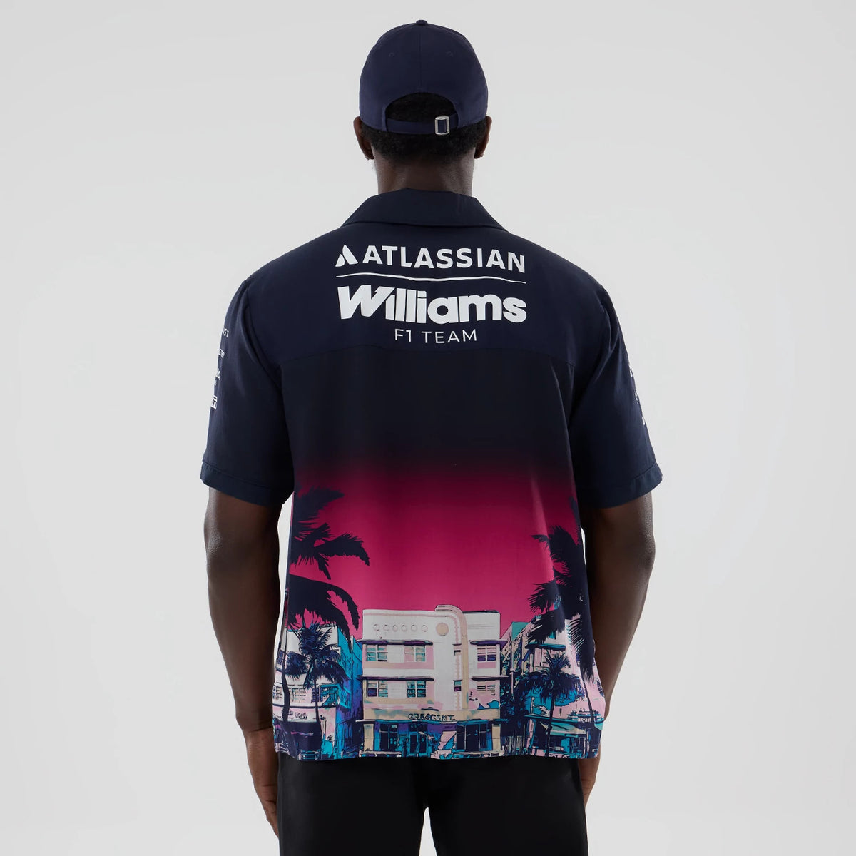 New Era Williams Miami Navy Short Sleeve Shirt, back view on a model with a navy cap, showing white Atlassian and Williams F1 Team lettering across the upper back and a pink and navy Miami cityscape and palm tree graphic on the lower section.
