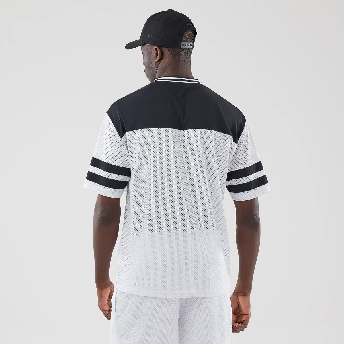 New Era Mesh White Oversized T-Shirt, back view on a model showing the black upper panel and collar, a white mesh body, and double black stripes on the cuffs of short white sleeves, styled with a black cap and white shorts.