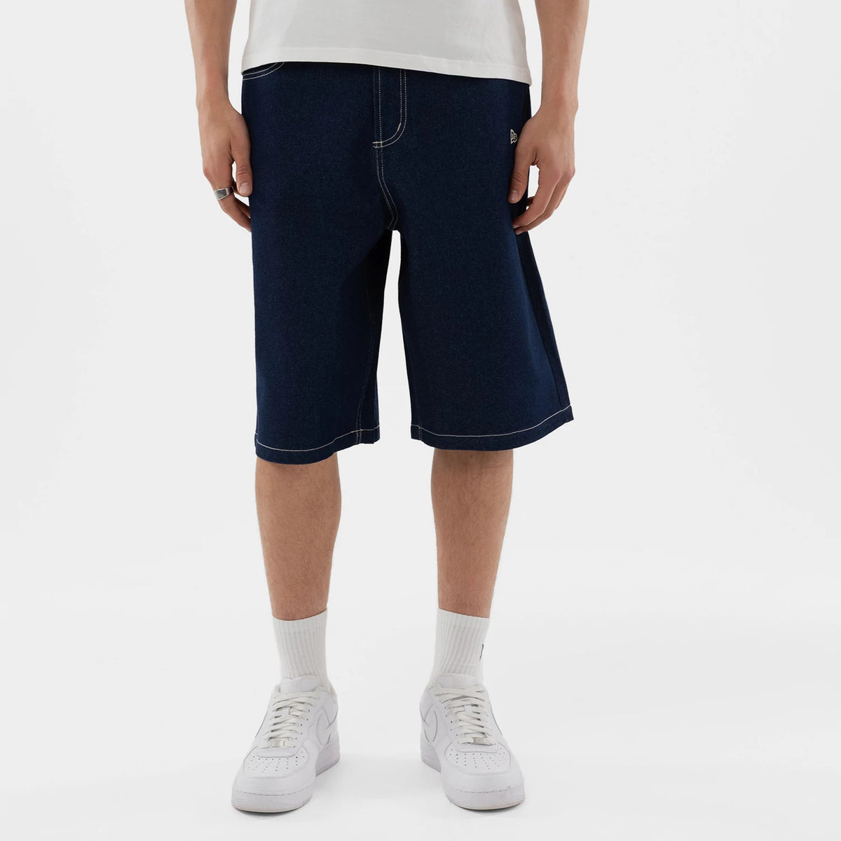 New Era New York Yankees MLB Jort Dark Blue Oversized Shorts, front view on a model showing the dark blue knee-length denim design with contrast stitching, an embroidered New Era logo on the left pocket and a relaxed fit above white trainers.