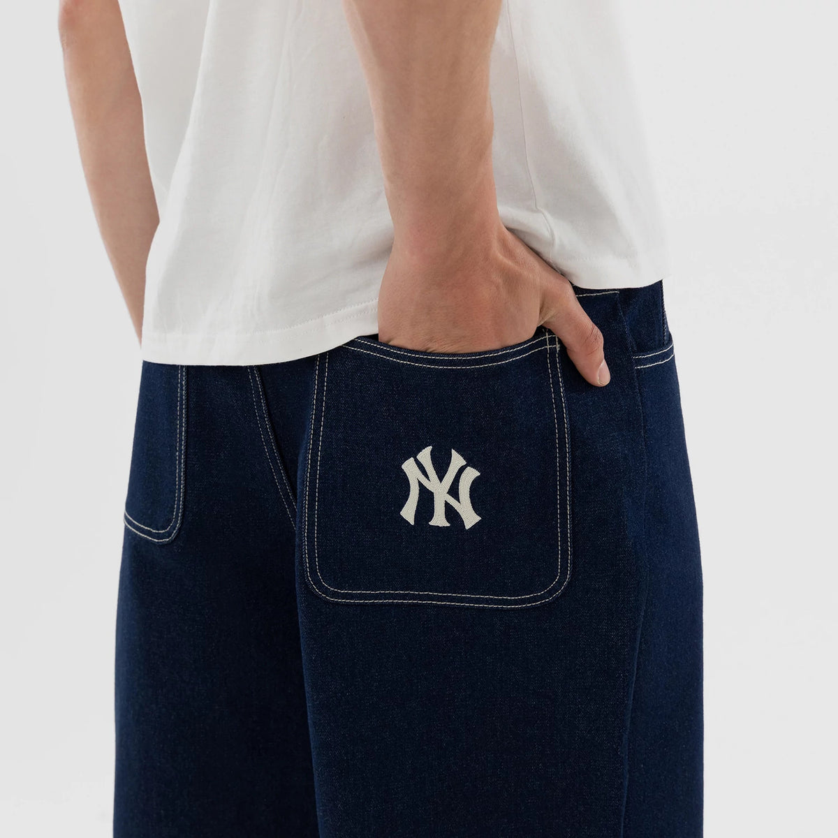 New Era New York Yankees MLB Jort Dark Blue Oversized Shorts, side view showing the dark blue fabric with contrasting white stitching, an embroidered white New York Yankees logo on the back pocket, and a casual styling with a white shirt tucked in.