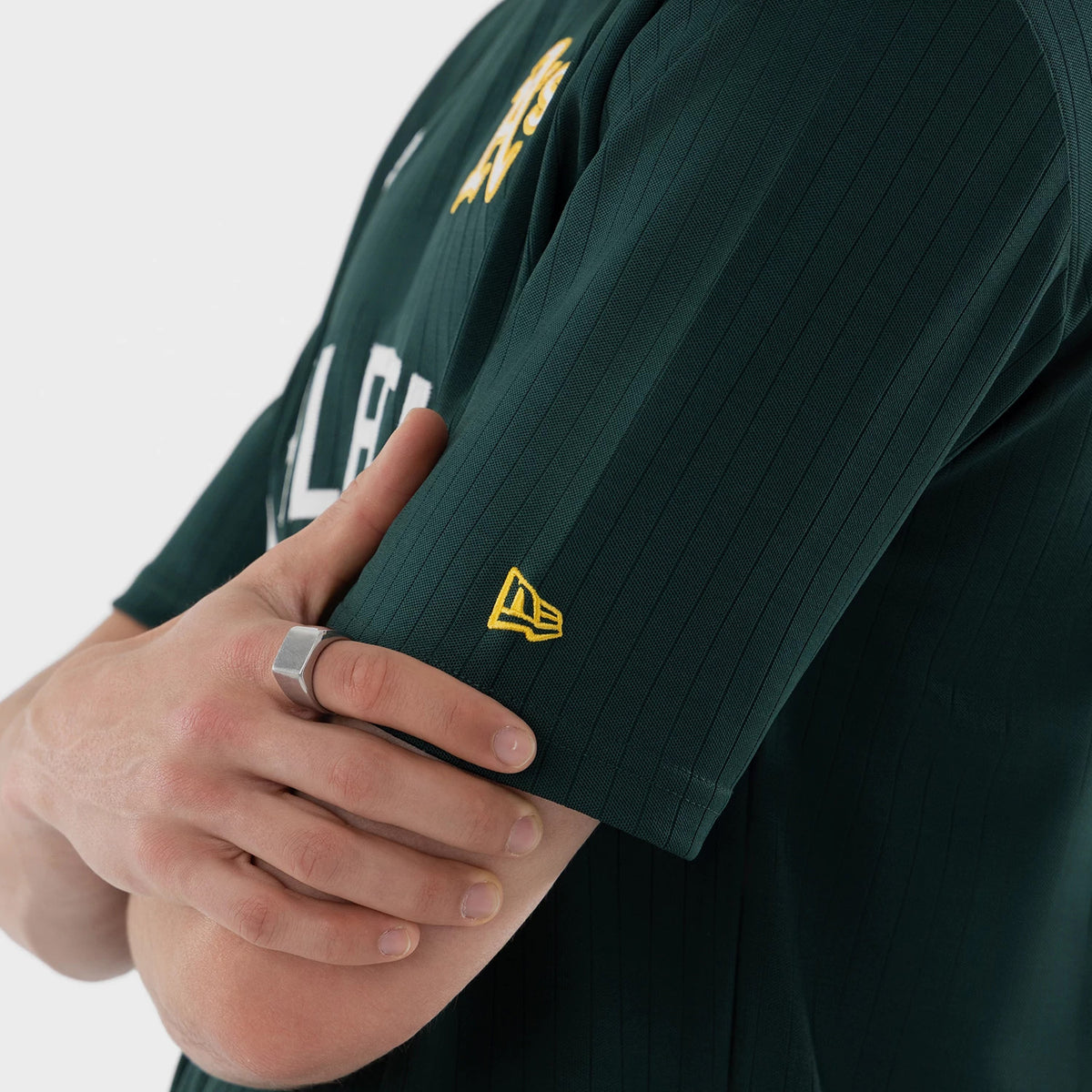 New Era Athletics MLB Dark Green Oversized T-Shirt, side view showing the dark green fabric with vertical stripes, a yellow embroidered New Era logo on the sleeve, and A’s lettering on the front worn by a model.