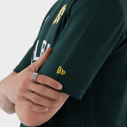 New Era Athletics MLB Dark Green Oversized T-Shirt, side view showing the dark green fabric with vertical stripes, a yellow embroidered New Era logo on the sleeve, and A’s lettering on the front worn by a model.