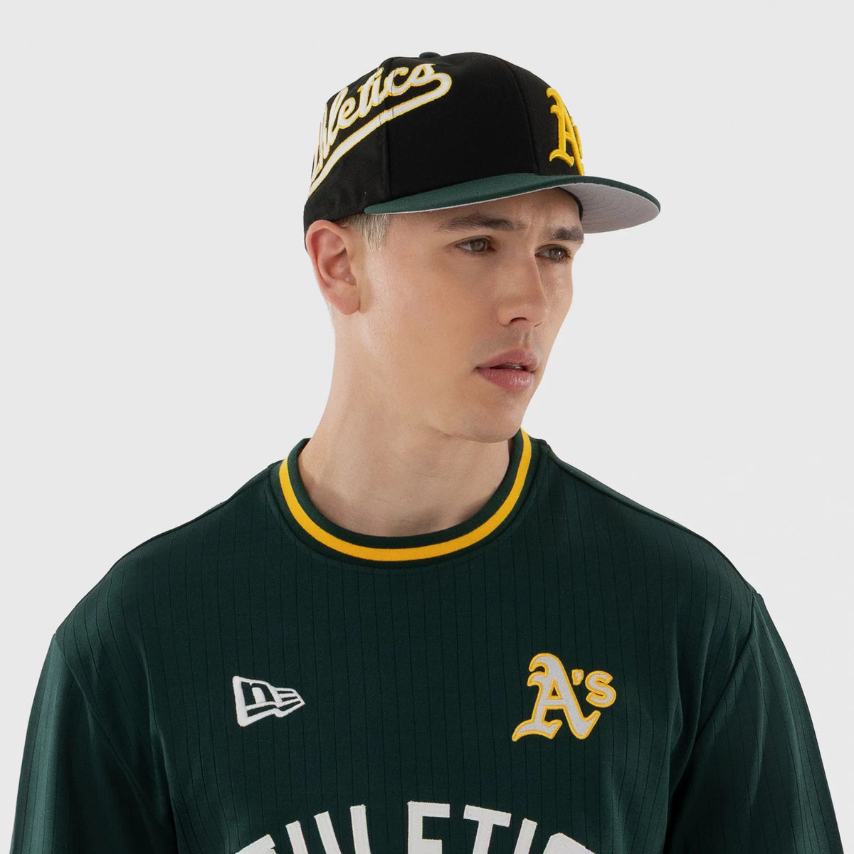 New Era Athletics MLB Dark Green Oversized T-Shirt, front view on a model styled with a matching cap, featuring the white New Era logo on the chest, A's in gold lettering on the left side, and ATHLETICS across the front.