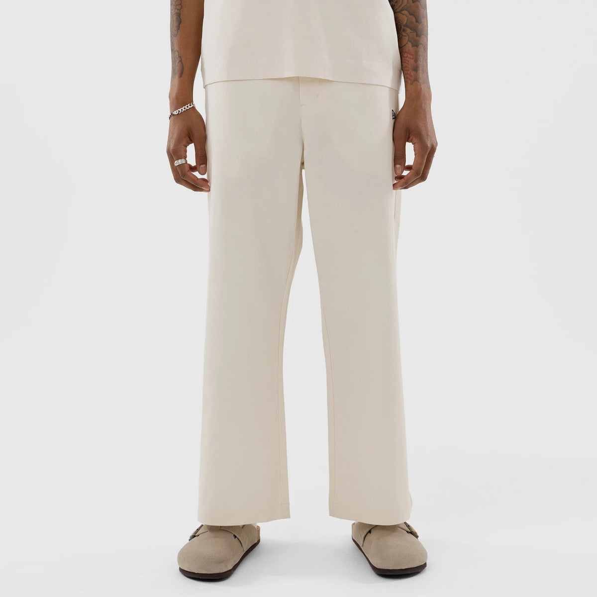 New Era New York Yankees MLB Cream Woven Chinos, front view on a model with loose-fitting legs and a straight cut, showcasing the New Era flag logo on the right pocket and the clean hemline falling over beige slip-on shoes.