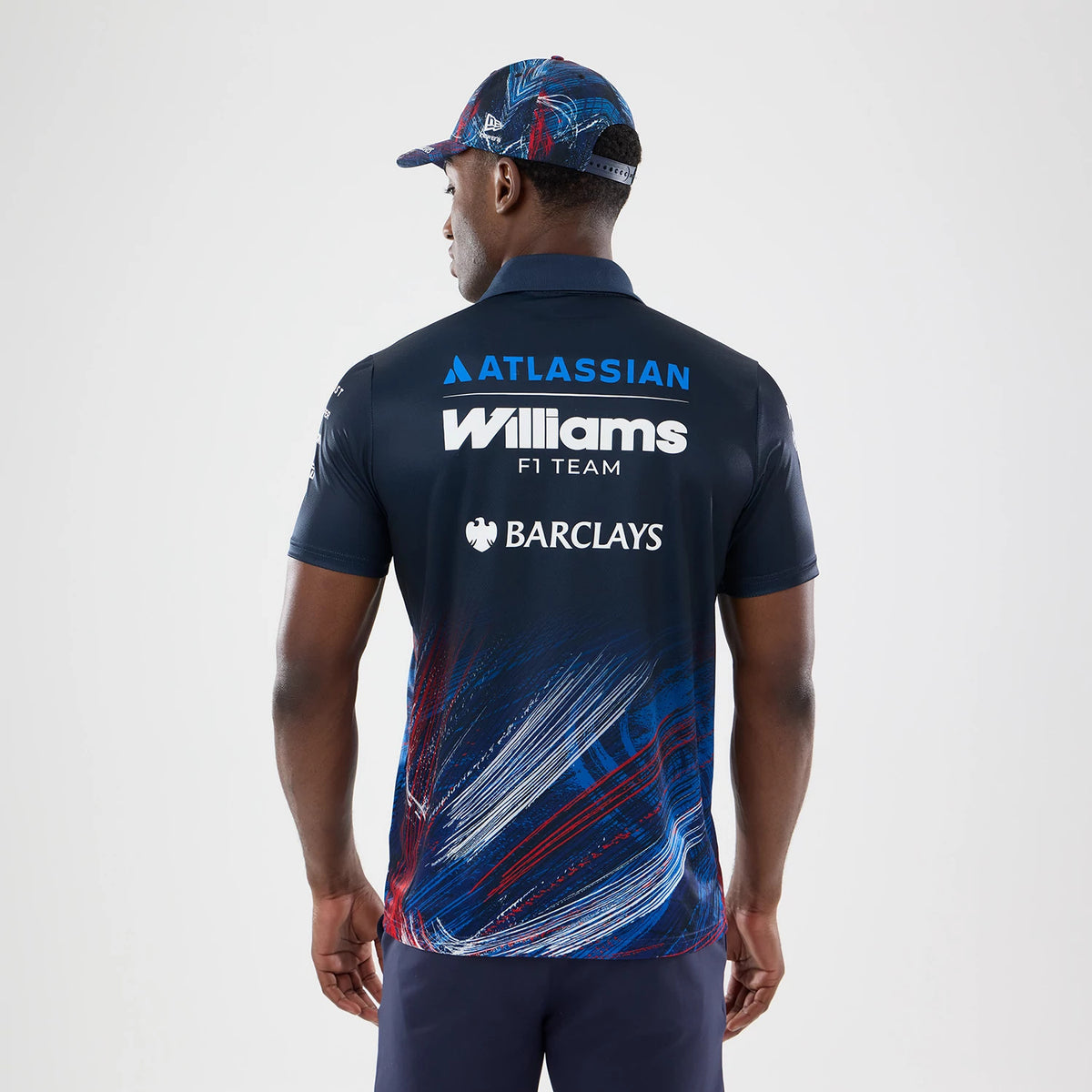 New Era Williams GB Race Special Navy Tech Polo T-Shirt, back view on a model with a coordinating cap, displaying prominent Atlassian, Williams F1 Team, and Barclays logos centred on the upper back, with a abstract red and blue graphic at the bottom.