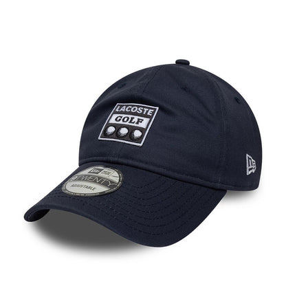 Lacoste X New Era Dark Blue 9TWENTY Adjustable Cap, three-quarter view showing a white embroidered Lacoste Golf badge with three golf balls centred on the front panel, dark blue curved visor with 9TWENTY size sticker.
