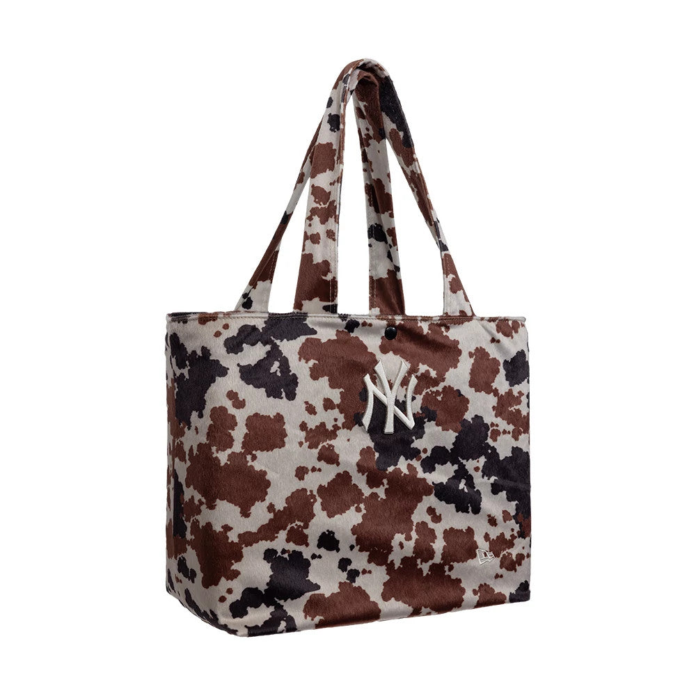 New Era New York Yankees MLB Cow Print Tote Bag, three-quarter view displaying a brown and black cow print pattern, two shoulder straps, an embroidered white NY logo on the front, and a small New Era logo near the bottom edge.