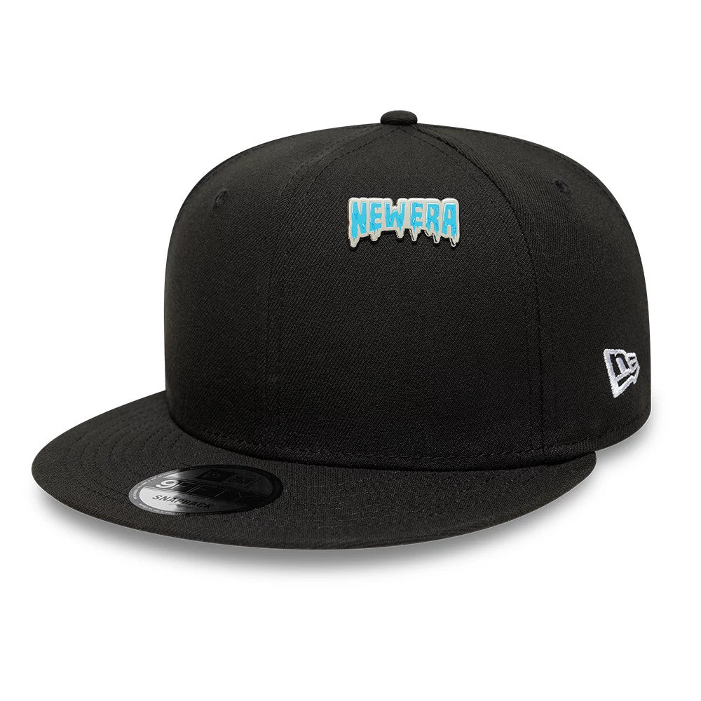 New Era Drip Script Silver Pin Badge, front view showing a black cap with a raised blue Drip Script pin badge centred on the crown, an embroidered New Era logo on the side and a flat black visor beneath the crown.