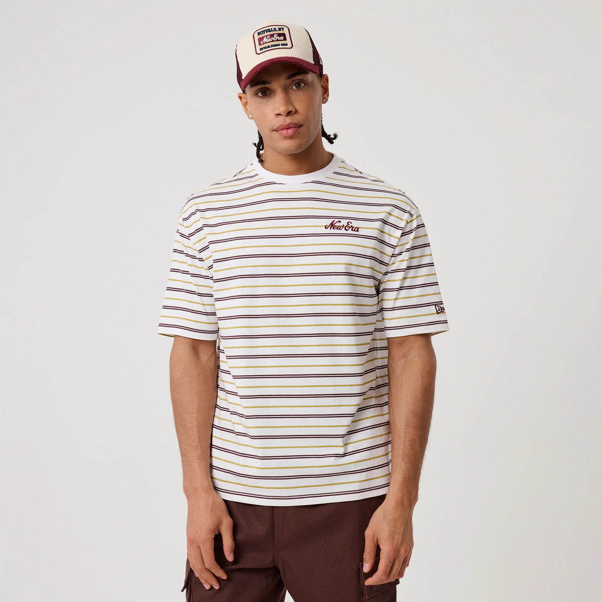 New Era Stripe White T-Shirt, front view on a model styled with a cap, showing horizontal stripes in brown, yellow, and purple, an embroidered New Era logo on the chest, and another embroidered logo on the left sleeve.
