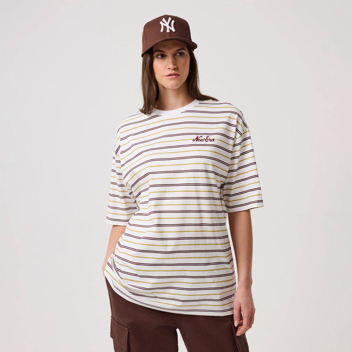 New Era Stripe White T-Shirt, front view on-model showing a relaxed fit with horizontal stripes in white, burgundy and gold, and an embroidered New Era logo on the left chest, paired with brown trousers and a brown cap.
