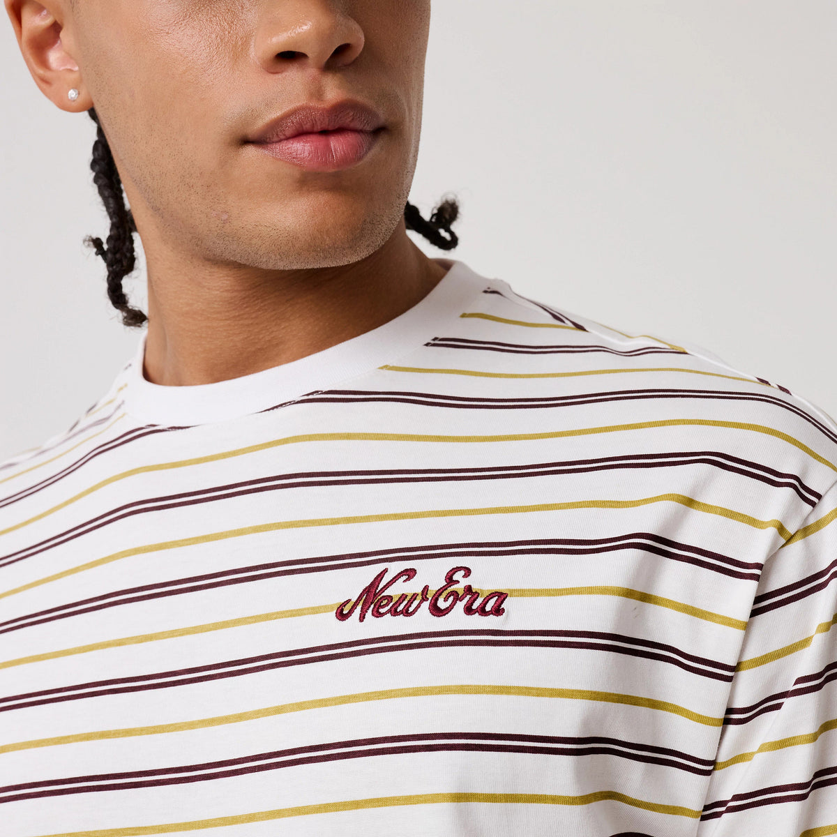 New Era Stripe White T-Shirt, front view on a model featuring horizontal brown and yellow stripes, a burgundy embroidered New Era logo on the left chest, and a classic crew neck design.