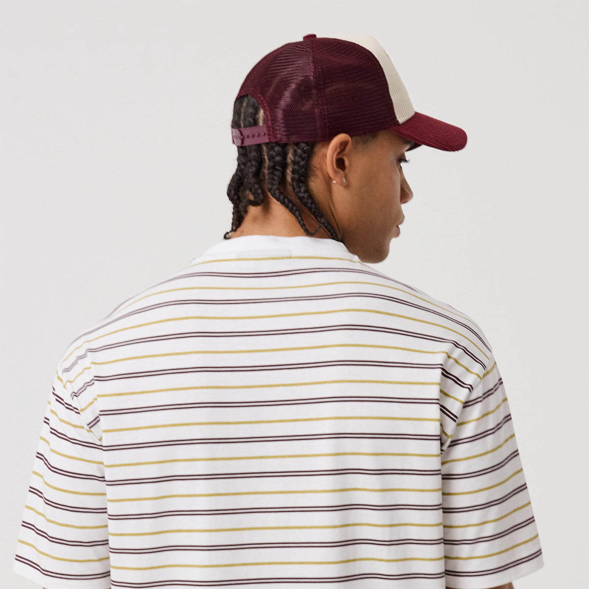 New Era Stripe White T-Shirt, back view on-model showing the white shirt with horizontal stripes in shades of black, yellow, and burgundy, styled with a burgundy and beige cap worn backwards, highlighting the casual, streetwear look.