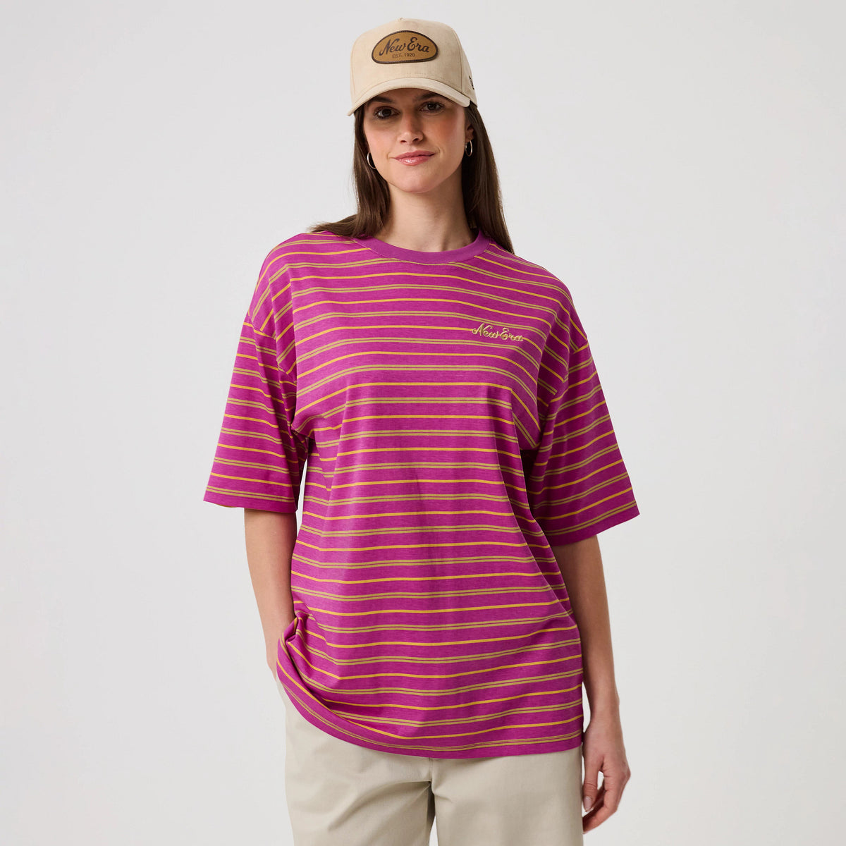New Era Stripe Bright Purple T-Shirt, front view on-model showcasing horizontal yellow stripes across the bright purple fabric, complemented by an embroidered New Era logo on the left chest, styled with beige trousers and a beige cap.