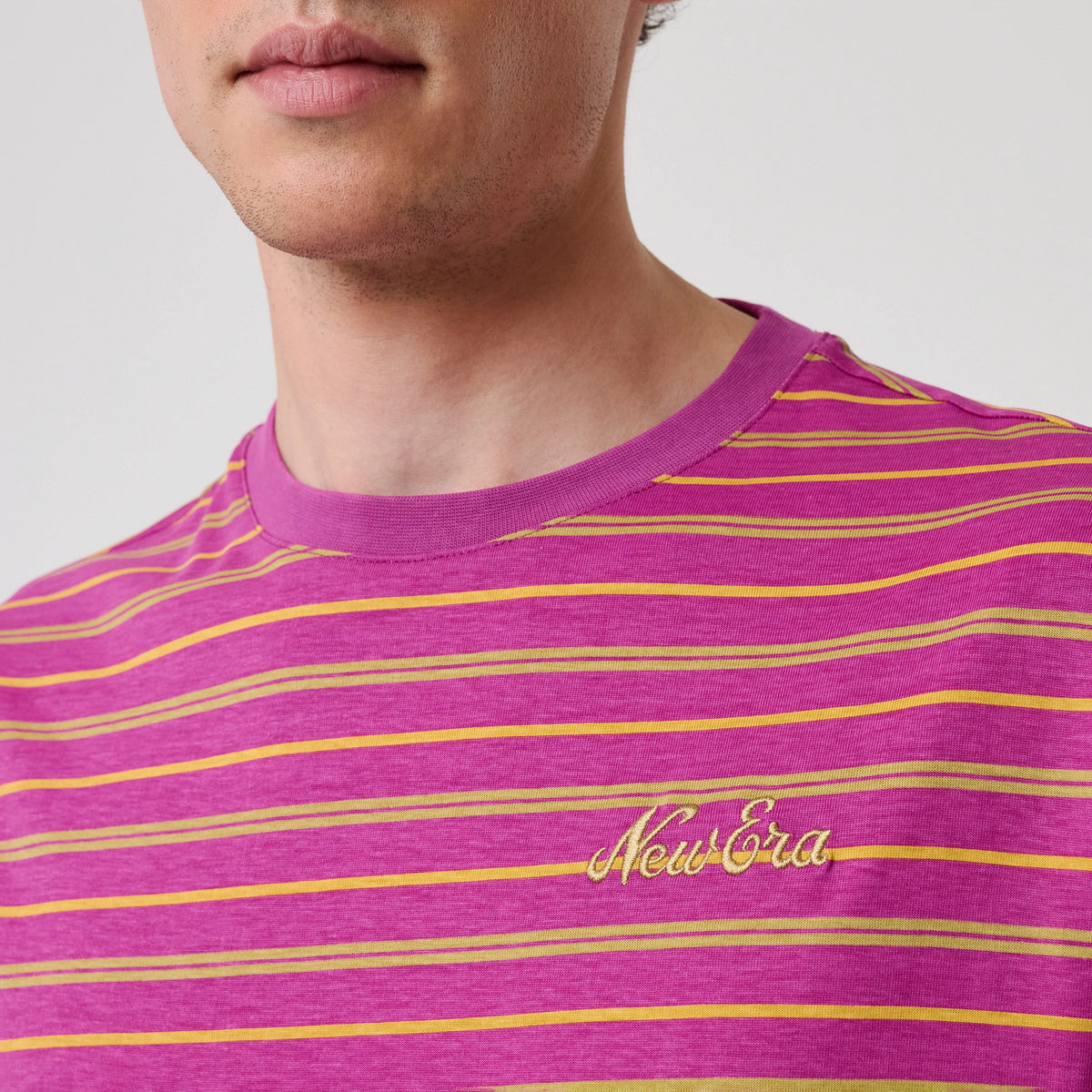 New Era Stripe Bright Purple T-Shirt, front view on a model highlighting the purple fabric with horizontal yellow and orange stripes, and an embroidered New Era logo in yellow on the left chest near the purple neckline.