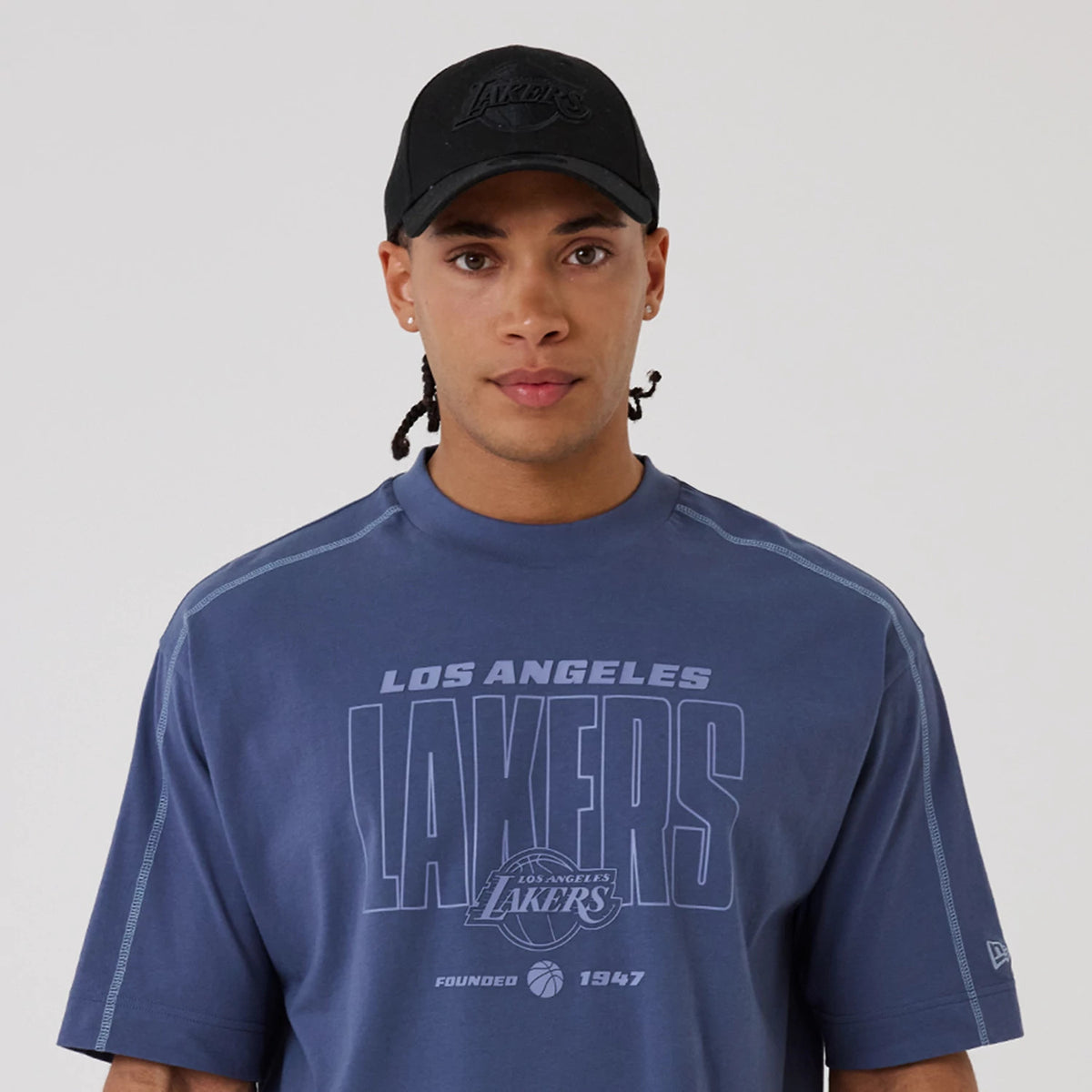 New Era LA Lakers NBA Graphic Dark Blue Boxy T-Shirt, front view on a model wearing a black cap, featuring large Lakers text across the chest, Los Angeles script above it, the team logo beneath, and additional lettering that reads 'Founded 1947'.