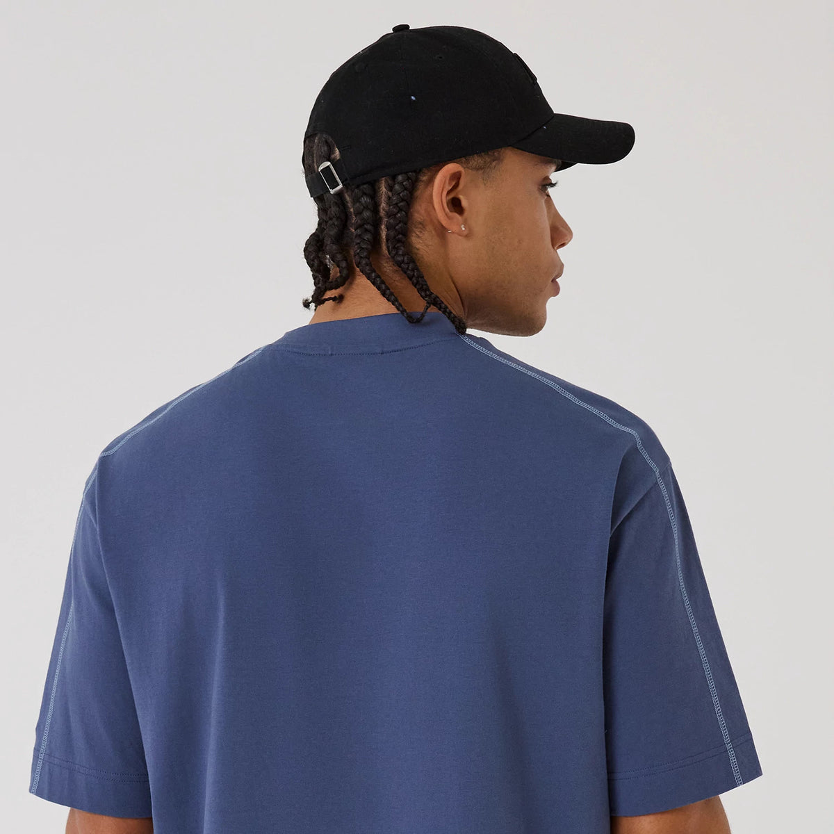 New Era LA Lakers NBA Graphic Dark Blue Boxy T-Shirt, back view on-model highlighting the plain dark blue fabric with stitching along the shoulders and sleeves, complemented by a black cap featuring an embossed New Era logo.