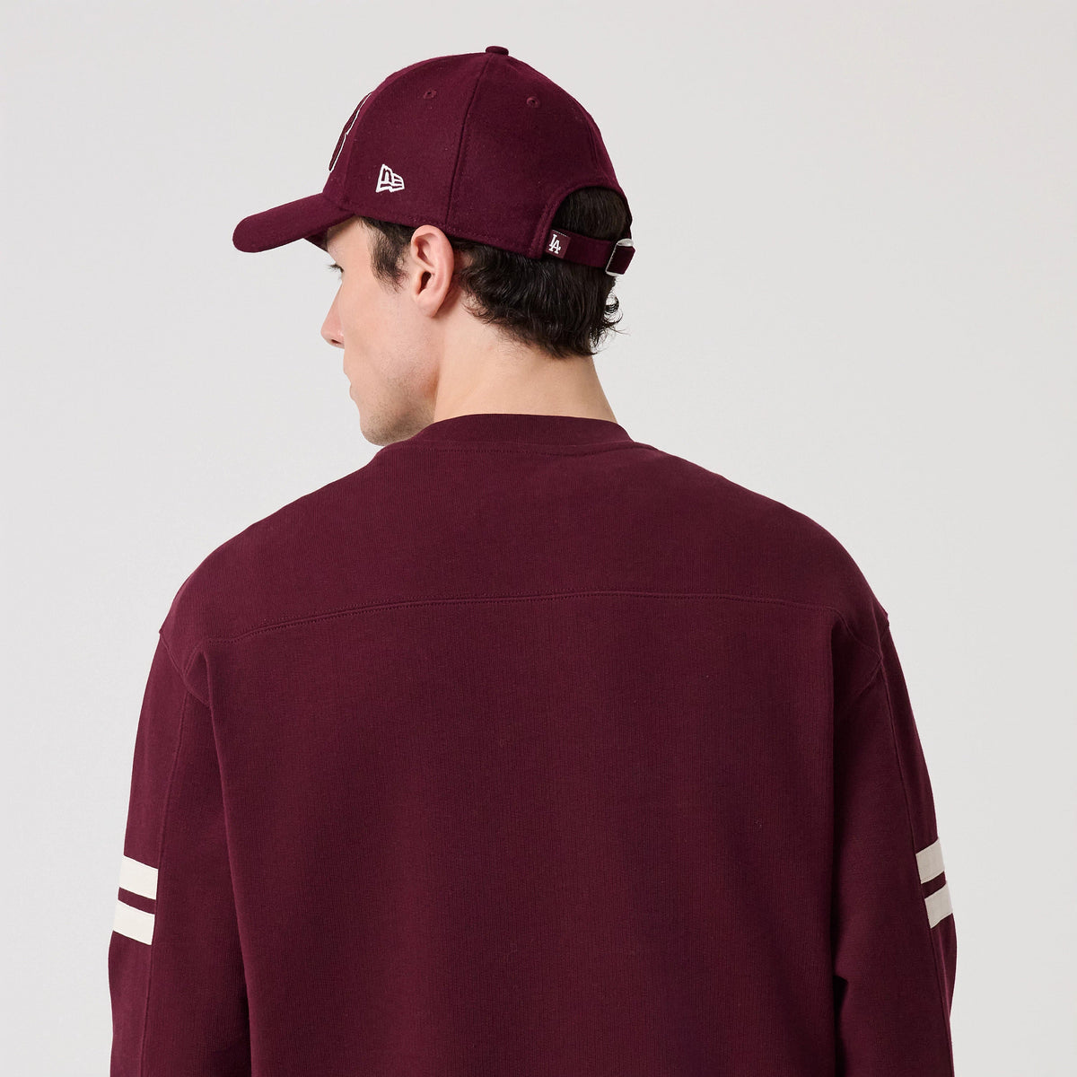 New Era LA Dodgers MLB Arch Graphic Dark Purple Crew Neck Sweater, back view on a model wearing a matching dark purple cap, showing white stripe on the sleeves and a relaxed fit in dark purple fabric.