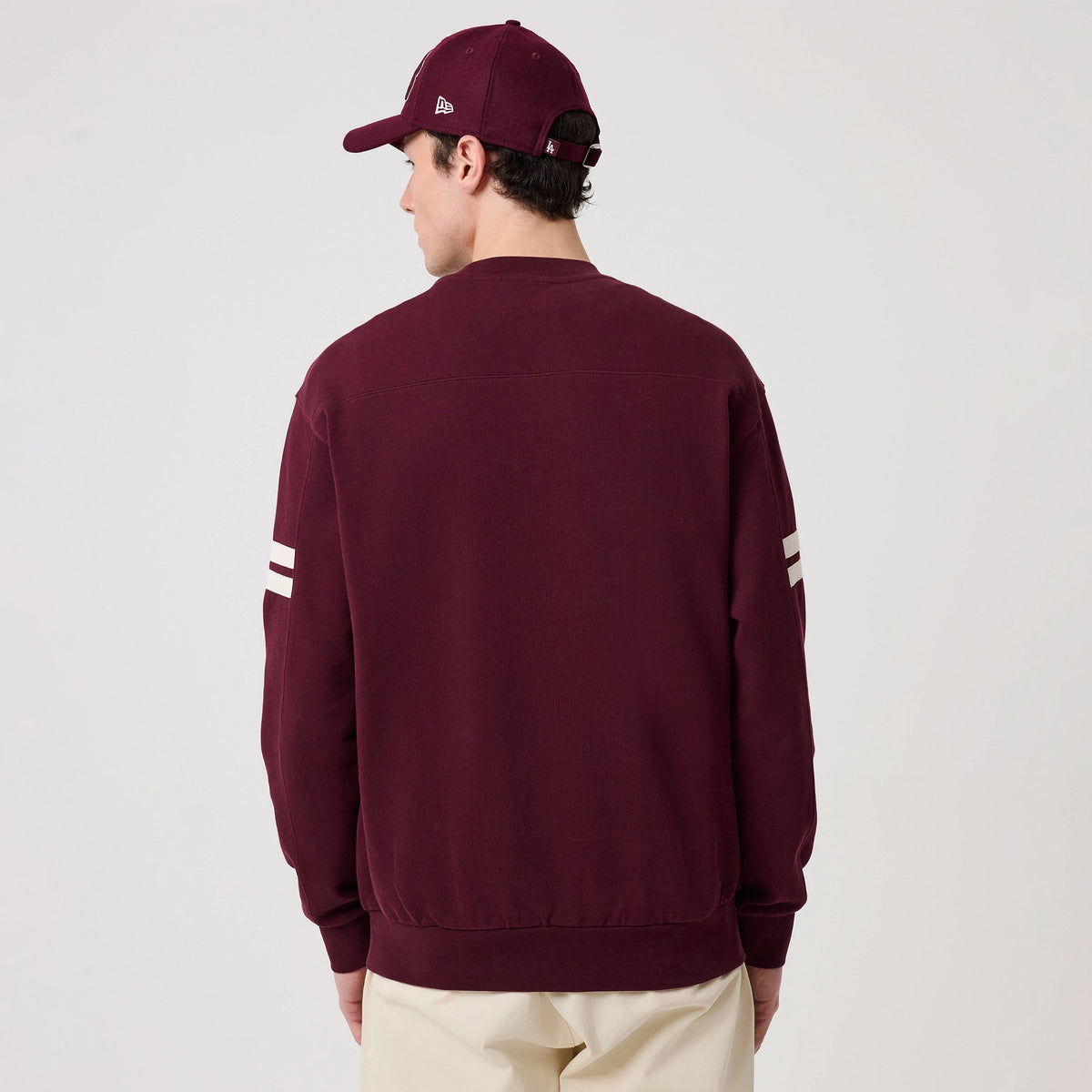 New Era LA Dodgers MLB Arch Graphic Dark Purple Crew Neck Sweater, back view on a model with a matching dark purple cap, showing two white stripes on each sleeve and a ribbed waistband, highlighting the sweater's relaxed fit.