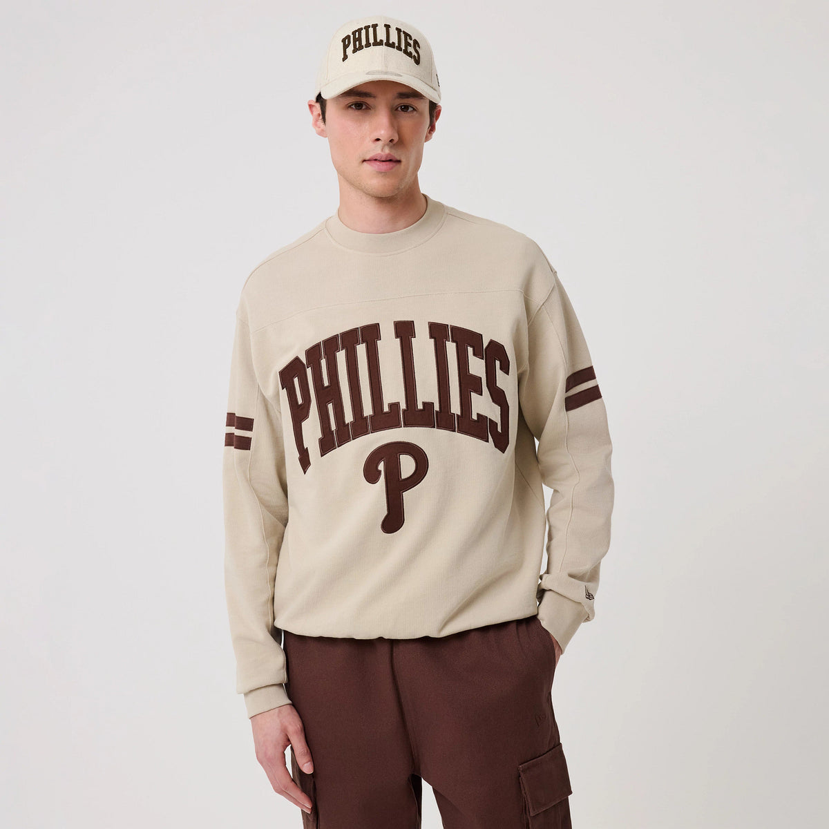 New Era Philadelphia Phillies MLB Arch Graphic Light Beige Crew Neck Sweater, front view on a model wearing a matching cap, featuring large brown Phillies lettering and logo on the chest, with double stripes on the sleeves and a relaxed fit.