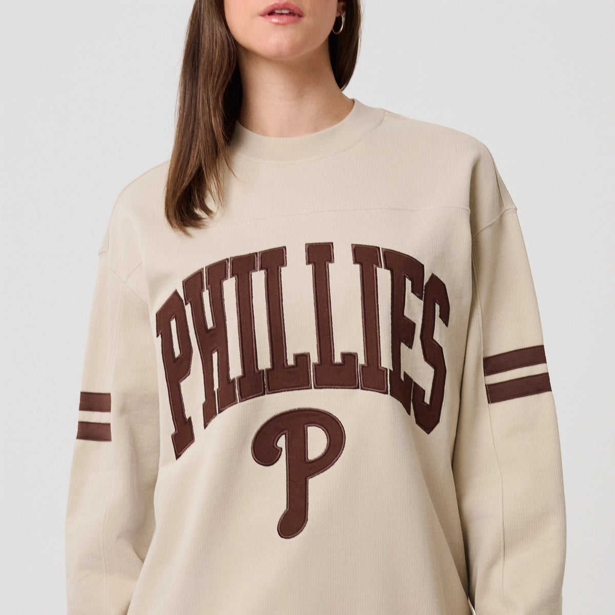 New Era Philadelphia Phillies MLB Arch Graphic Light Beige Crew Neck Sweater, front view on a model showcasing the brown PHILLIES text in arch embroidery across the chest, with matching double-stripe on both sleeves.