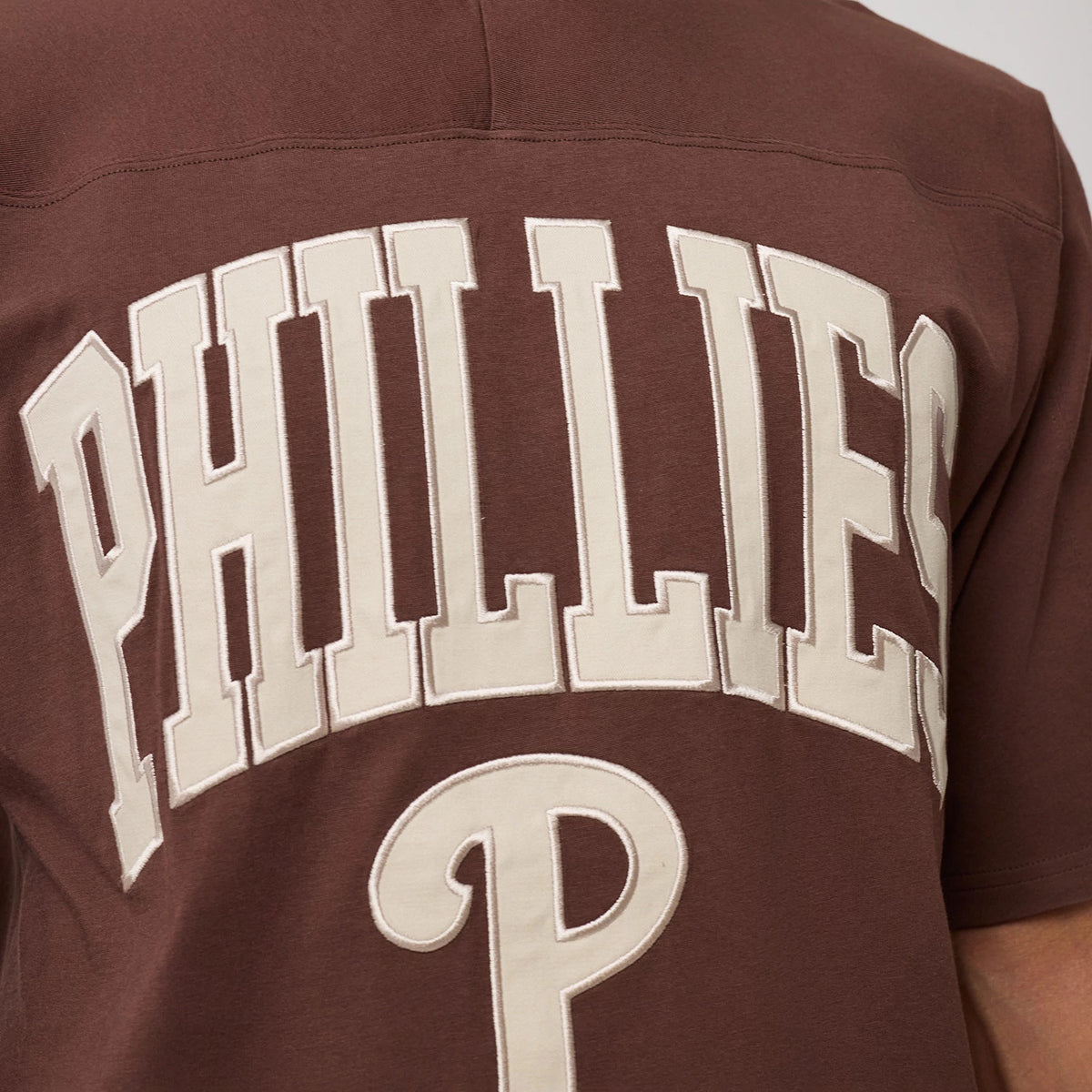 New Era Philadelphia Phillies MLB Arch Graphic Brown Oversized T-Shirt, back view showcasing prominent PHILLIES lettering in white arched across the upper back, with a large embroidered P logo below, set against a rich brown fabric backdrop.