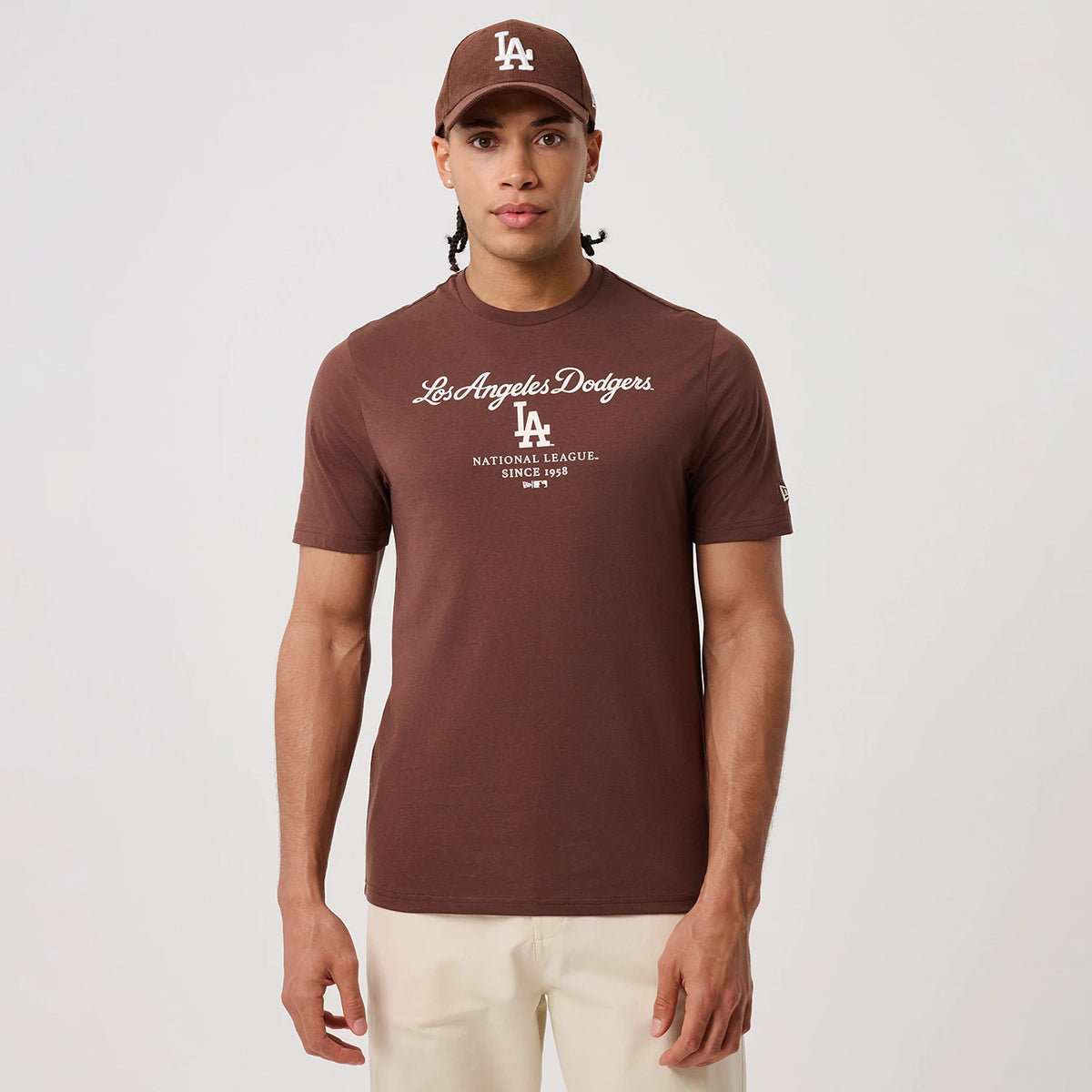 New Era LA Dodgers MLB Linear Brown T-Shirt, front view on a model with a matching brown cap, showcasing white embroidered Los Angeles Dodgers script across the chest, LA logo beneath, and New Era flag on the left sleeve.