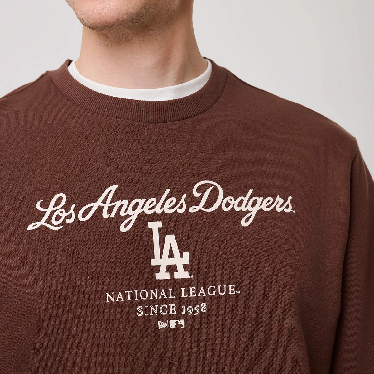 New Era LA Dodgers MLB Linear Brown Crew Neck Sweater, front view on a model featuring white Los Angeles Dodgers script embroidered across the chest, complemented by the LA logo beneath and MLB branding near the hem, with ribbed crew neck collar.