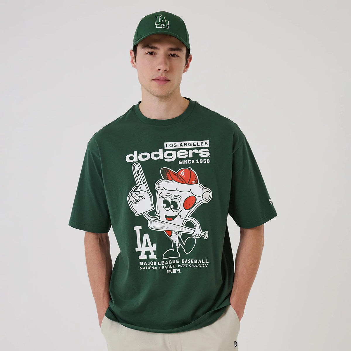 New Era LA Dodgers MLB Food Graphic Dark Green Oversized T-Shirt, front view on a model with a matching green cap, featuring a graphic of a cartoon pizza with a bat and glove, and white Los Angeles Dodgers lettering across the chest.