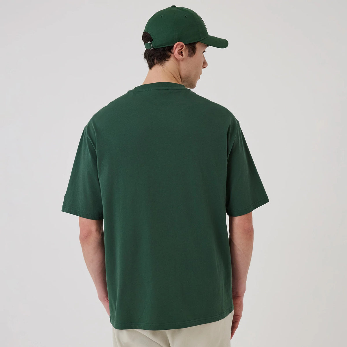 New Era LA Dodgers MLB Food Graphic Dark Green Oversized T-Shirt, back view on a model with a matching dark green cap, showing the plain dark green back of the shirt with loose short sleeves and a straight hemline, styled with light trousers.
