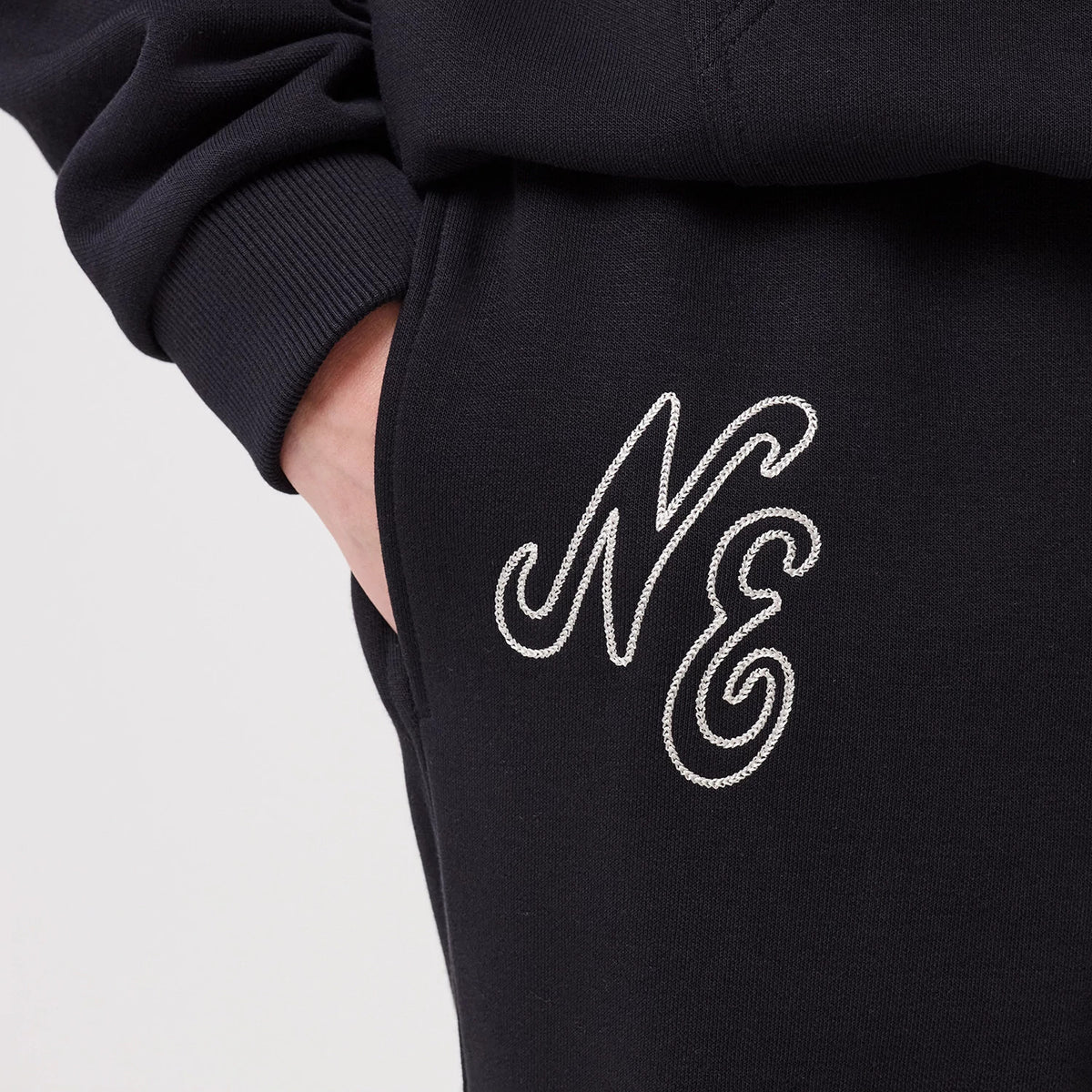 New Era Navy Straight Leg Joggers, close-up showing the embroidered script NE logo in white on the upper thigh, with a hand placed in the side pocket, and a ribbed cuff seen above the logo, highlighting the garment's textured material.