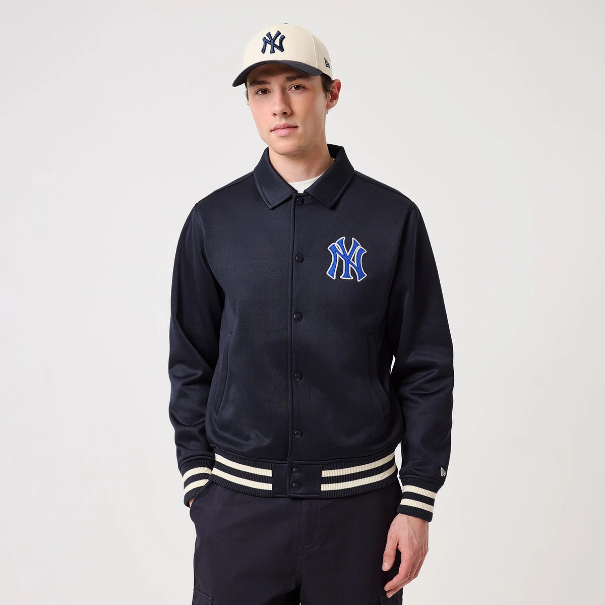 New Era New York Yankees MLB Navy Collared Jacket, front view on a model styled with a beige cap, featuring a prominent embroidered Yankees logo on the left chest, button-up closure, and ribbed cuffs and hem with white and navy stripes.