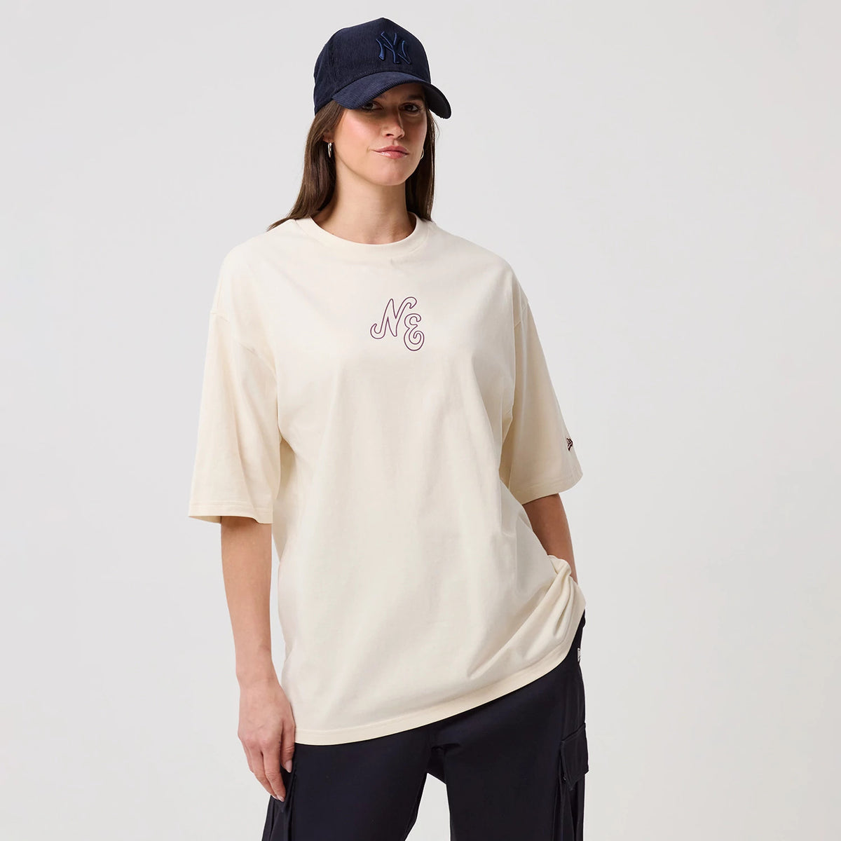 New Era Crest Graphic Open White Oversized T-Shirt, front view on-model showing the relaxed white fit with large NC graphic on the chest, short sleeves and a small embroidered New Era logo on the left sleeve, paired with a dark cap.