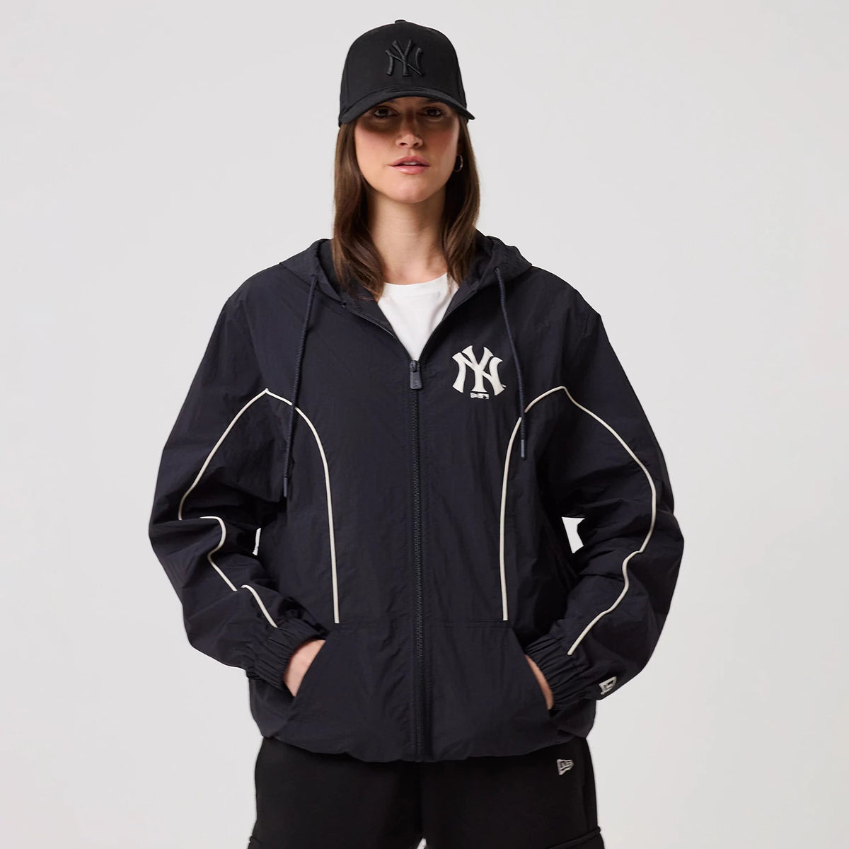 New Era New York Yankees MLB Piping Navy Track Jacket, front view on-model showing the navy jacket with white piping along the sleeves and sides, an embroidered team logo on the left chest, and the model styled in a matching black cap.