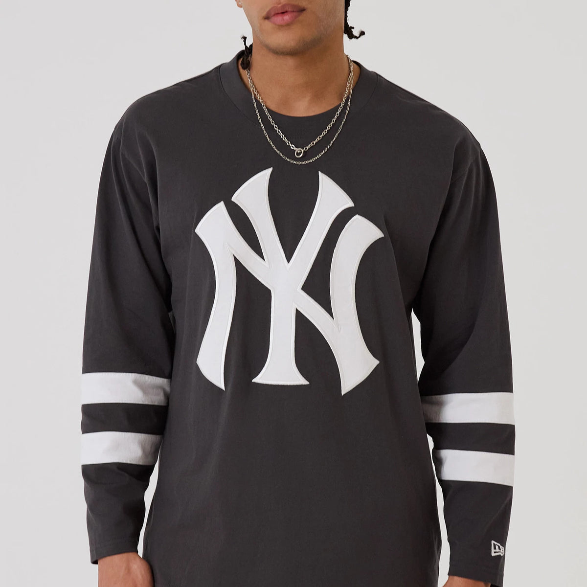 New Era New York Yankees MLB League Essential Charcoal Oversized Jersey, front view on a model wearing jewellery, displaying a large white printed Yankees logo on the chest and two white horizontal stripes on each sleeve near the cuffs.
