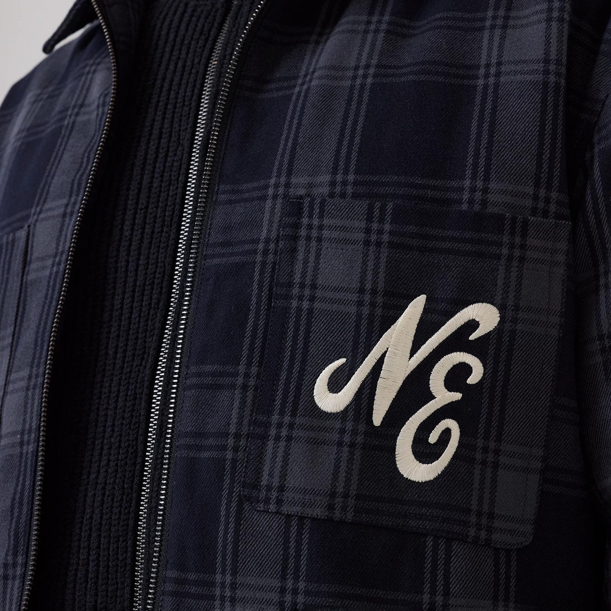 New Era Navy Shacket, close-up view showing the embroidered cream NE initials on the left chest pocket, a navy and grey plaid pattern, and contrasting exposed zip running along the front, layered over a textured black knit garment.