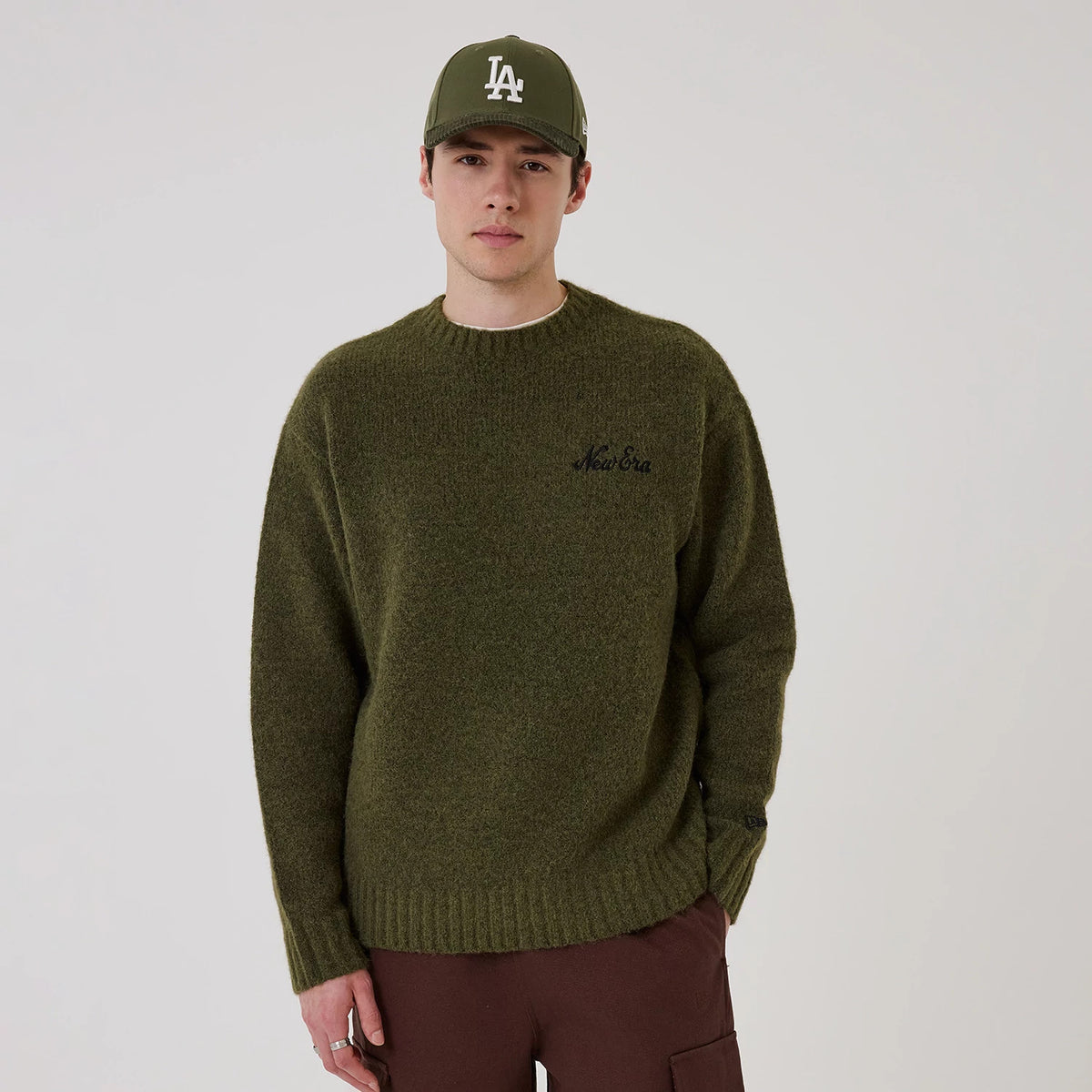 New Era Simple Knit Green Knitted Sweater, front view on a model wearing a green cap with a white LA logo, featuring a black embroidered New Era logo on the left chest and ribbed cuffs and hem.