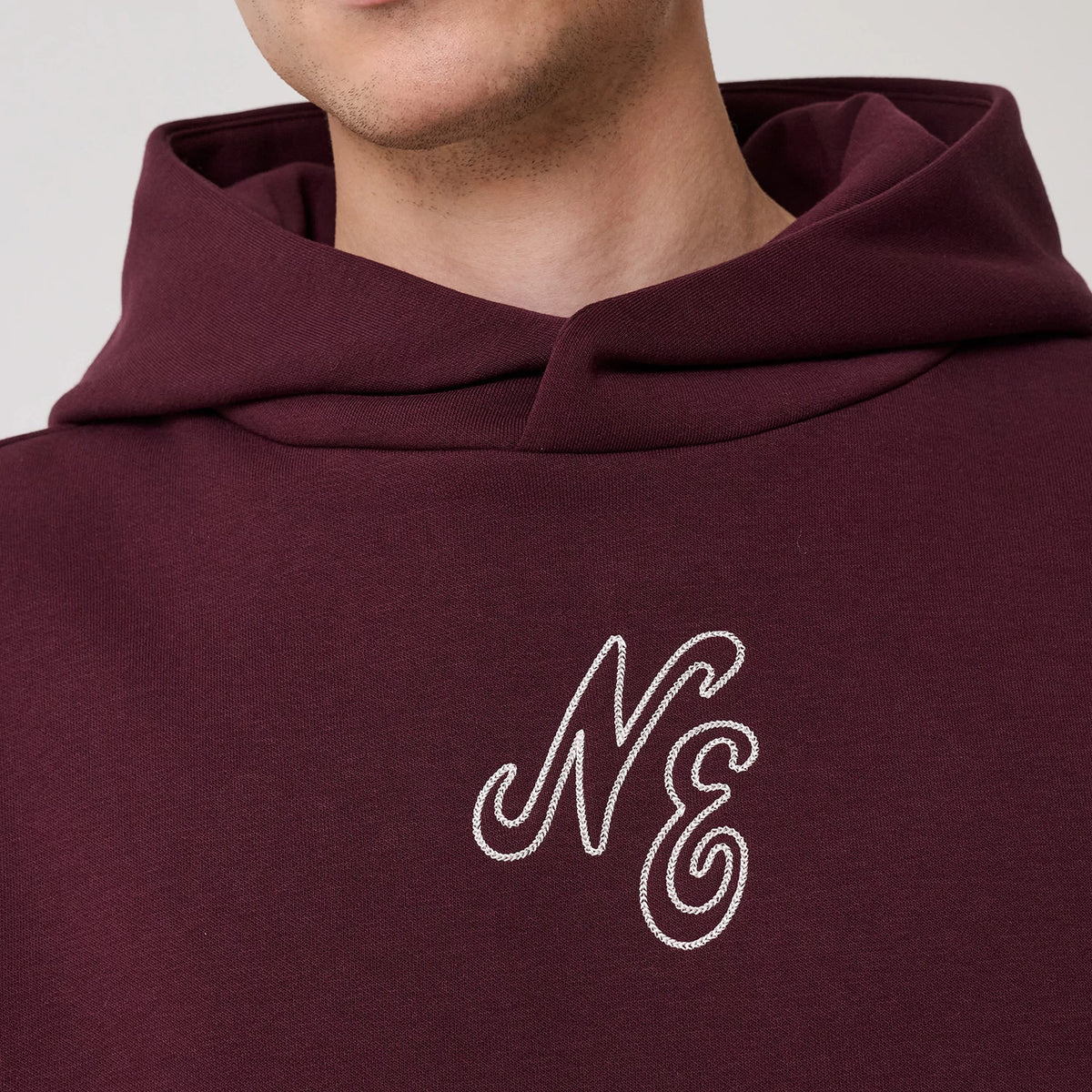New Era Crest Graphic Dark Purple Pullover Hoodie, front view showing a large hood and a prominently embroidered NE logo in white on the chest, with the dark purple fabric creating a contrast.